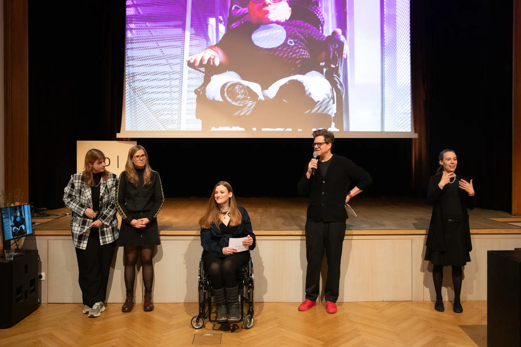 im Hintergrund eine Powerpoint mit einem Foto von Martin Habacher. Auf der B&uuml;hne spricht Stefan Wolner. Daneben sieht man v.l.n.r. Cornelia Ohnmacht, Hannah Wahl und die Moderatorin Barbara Sima Ruml.