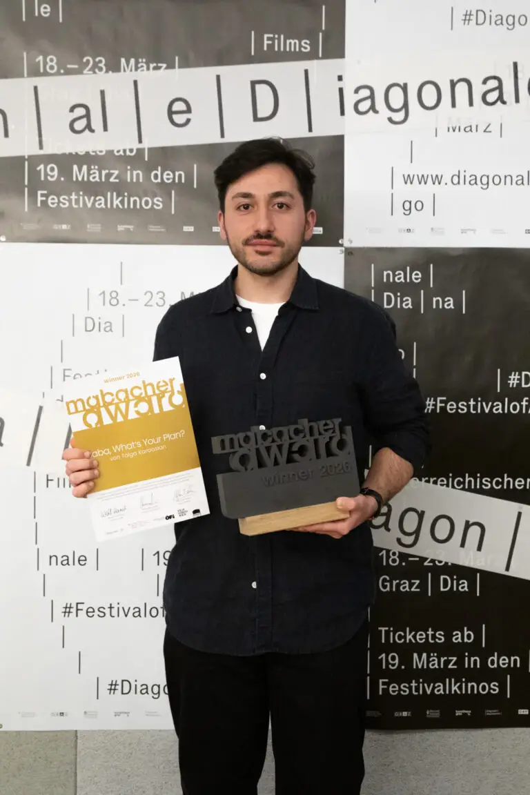Tolga Karaaslan. Ein junger Mann mit dunklen kurzen Haaren und Bart. Er trägt ein Hemd. Er hält eine Mabacher Award Urkunde und einen Preisskulptur in der Hand.