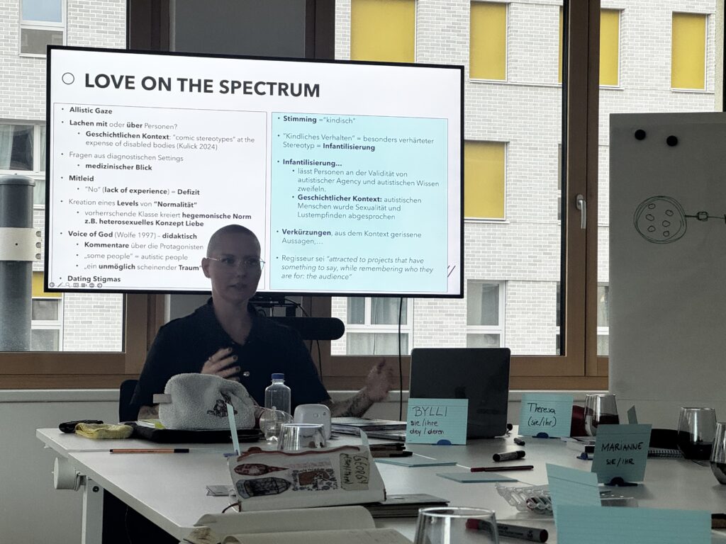 Sybille Bauer-Zierfuß sitz an einem Tisch. Dahinter sieht man einen Bildschirm mit Powerpoint.