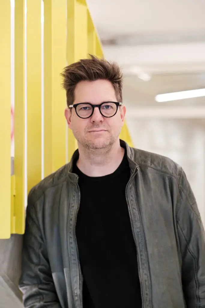 Stefan Wolner. Er trägt kurzes braunes Haar, eine schwarze Brille eine graue Lederjacke und blickt in die Kamera.